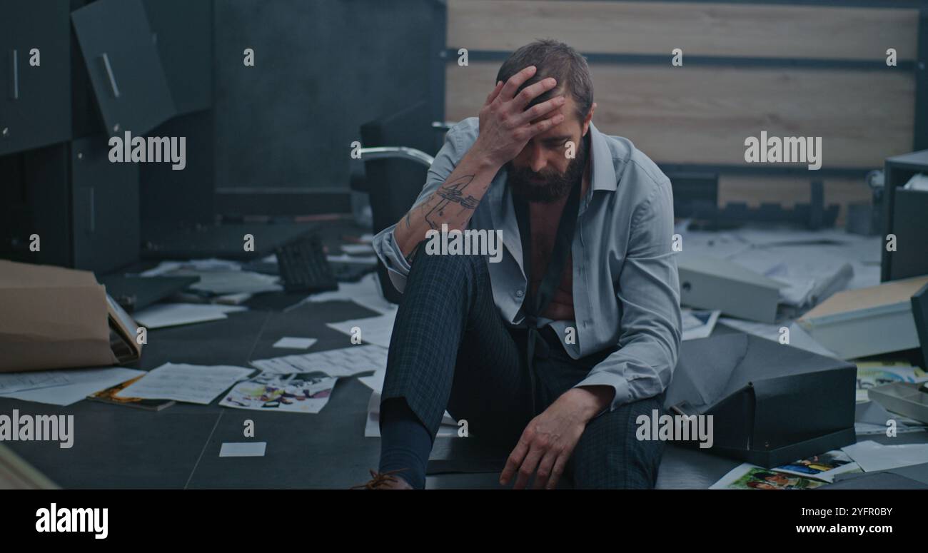 Nervous and stressed employee breathes heavily, sits down on floor after destroying office room due to firing. Frustrated businessman after failed business deal or receiving bad news. Financial crisis Stock Photo