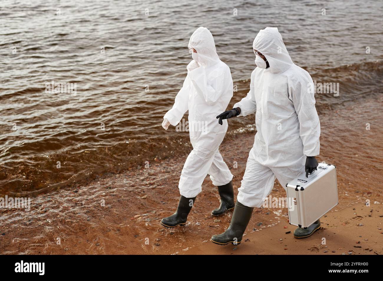 High angle view of two scientists wearing protective suits while walking along water and carrying toolkit case, copy space Stock Photo