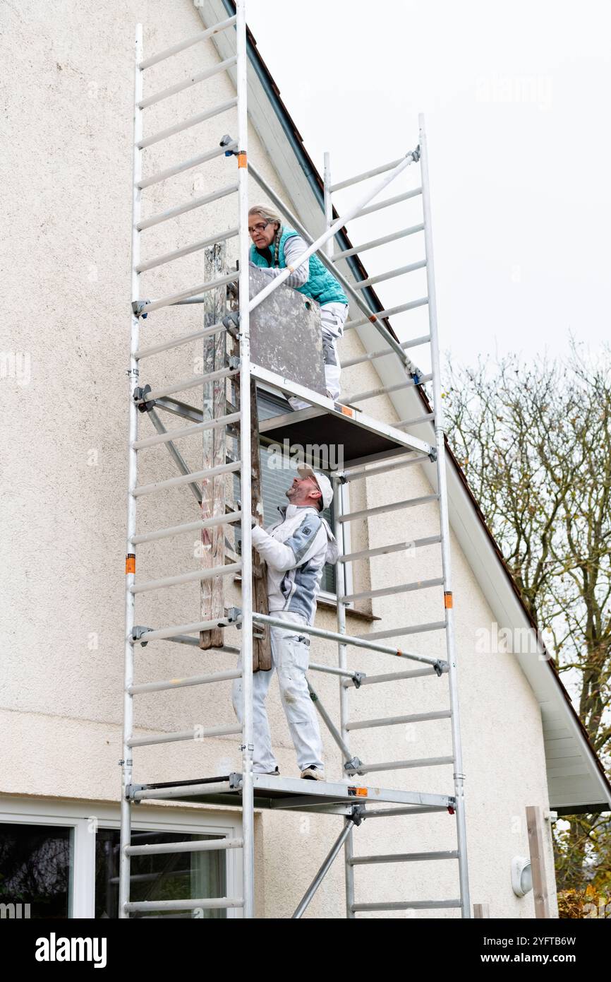 Gerüstbauerin, Malerin lässt sich vom Malerkollegen die Holzleiter nach oben auf das Gerüst reichen. mobiles Gerüst am Einfamilienhaus model released für redaktionelle Nutzung, Gerüstbauer *** Scaffolder, painter has a colleague hand her the wooden ladder up onto the scaffolding mobile scaffolding on a detached house model released for editorial use, scaffolder 20241105-DSC 8729 Stock Photo