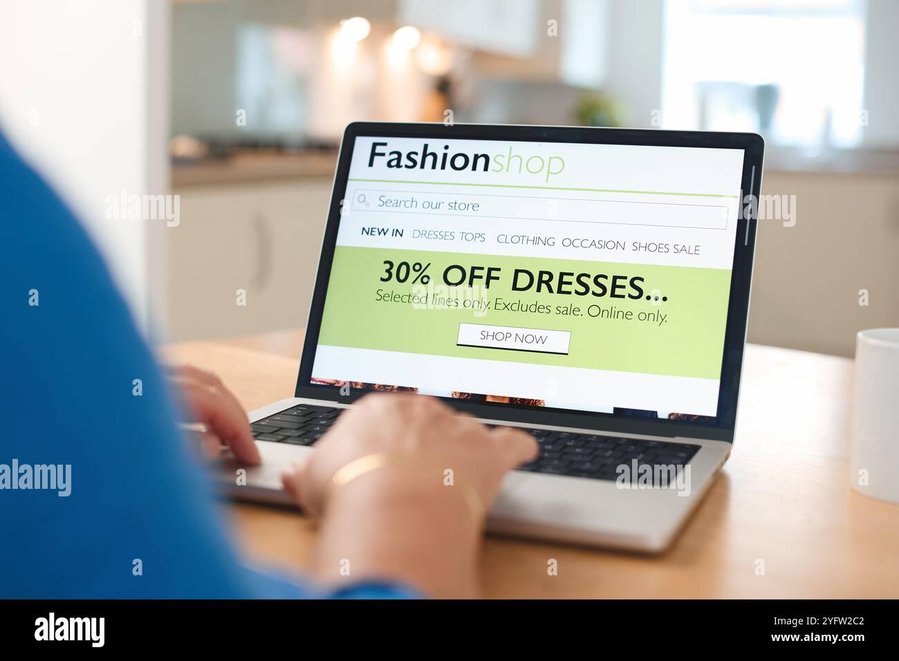 A person is using a laptop in a bright kitchen, engaged in online shopping. The screen shows a vibrant advertisement offering a discount on dresses, i Stock Photo