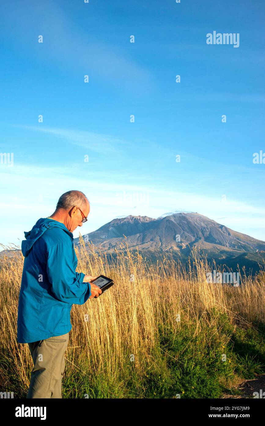 Adult senior male relaxes while reading a kindle book with Mount Saint Helensin the background. Northern Cascades, Washington State. USA Stock Photo