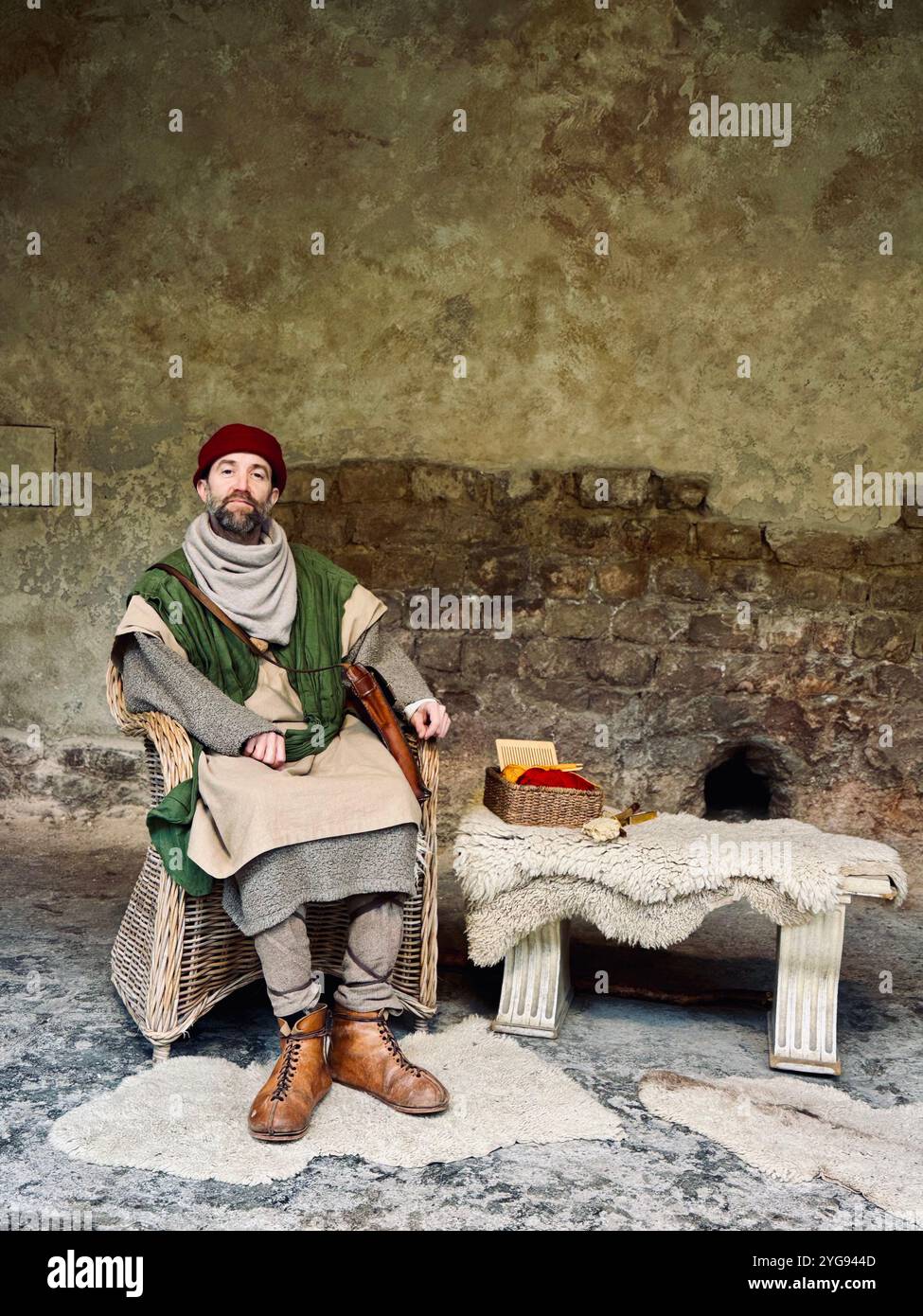 'A man dressed in historical attire sits in a wicker chair against a rustic stone wall, evoking a sense of ancient times.' - Smartphone Captured Stock Image