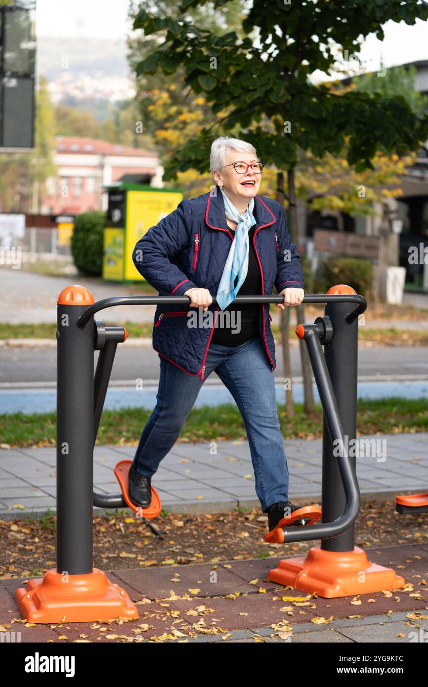 A mature woman in her seventies uses outdoor gym equipment to stay active in a peaceful autumn park setting. With a casual blue outfit and scarf, she Stock Photo