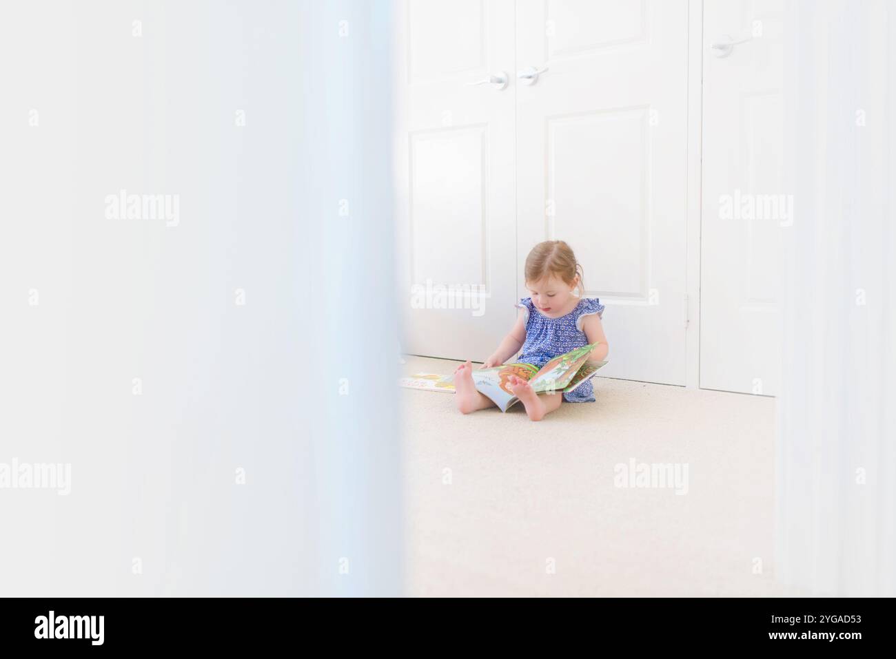 Young girl sitting on the floor in a bright room reading a picture book ...