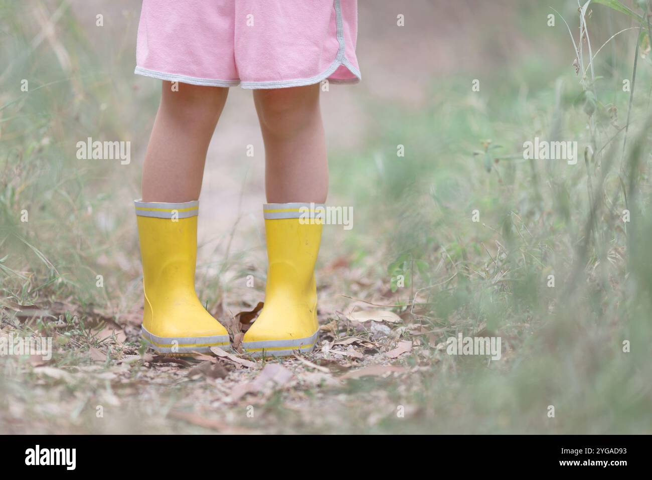 Child wearing yellow rubber boots and pink shorts standing on a path ...