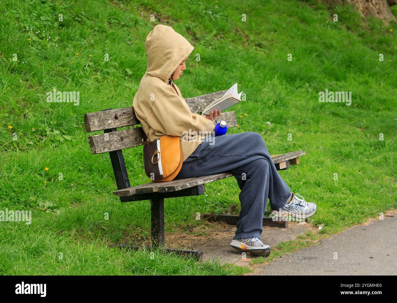 24 April 2024 A young female wearing a hoodie relaxing on a wooden bench while reading a book on the path alongside the River Avon in Bath Somerset En Stock Photo