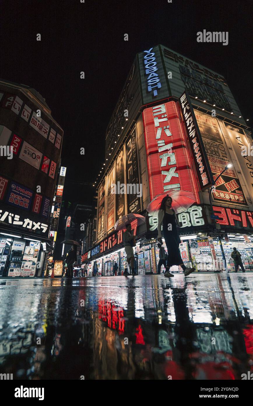 A woman with an umbrella walks past the Yodobashi Kamera shop in Shinjuku in the rain in the evening. The neon lights are reflected on the wet street Stock Photo