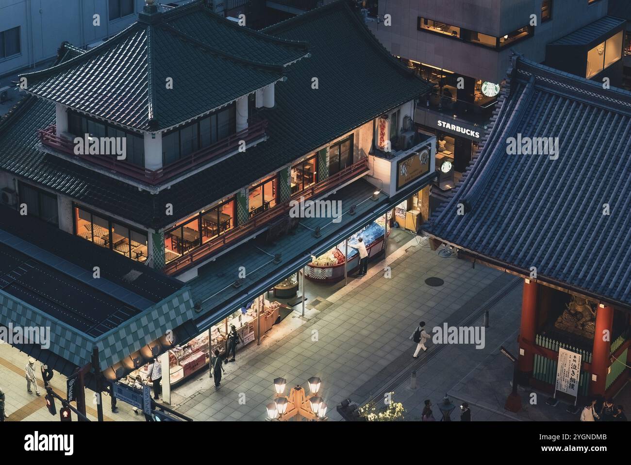 A woman walks past the sweetshop Tokiwado Kaminari-okoshi Honpo, Starbucks and the Kaminarimon entrance gate on the forecourt of Senso-ji. Senso-ji in Stock Photo