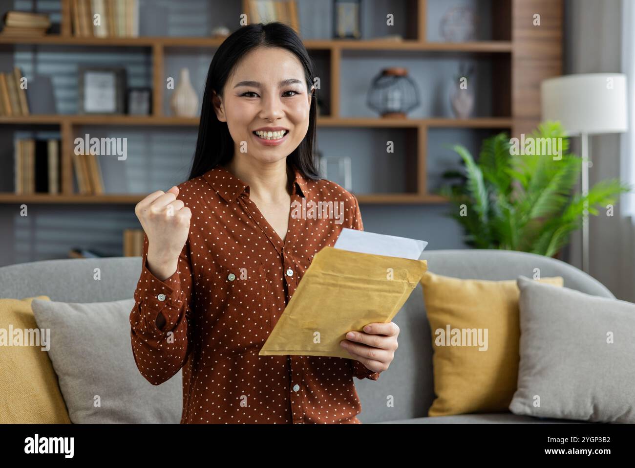 Asian woman expressing joy at home holding envelope in hand. Her happy and victorious expression represents success and positive news. Setting is cozy and comfortable. Stock Photo