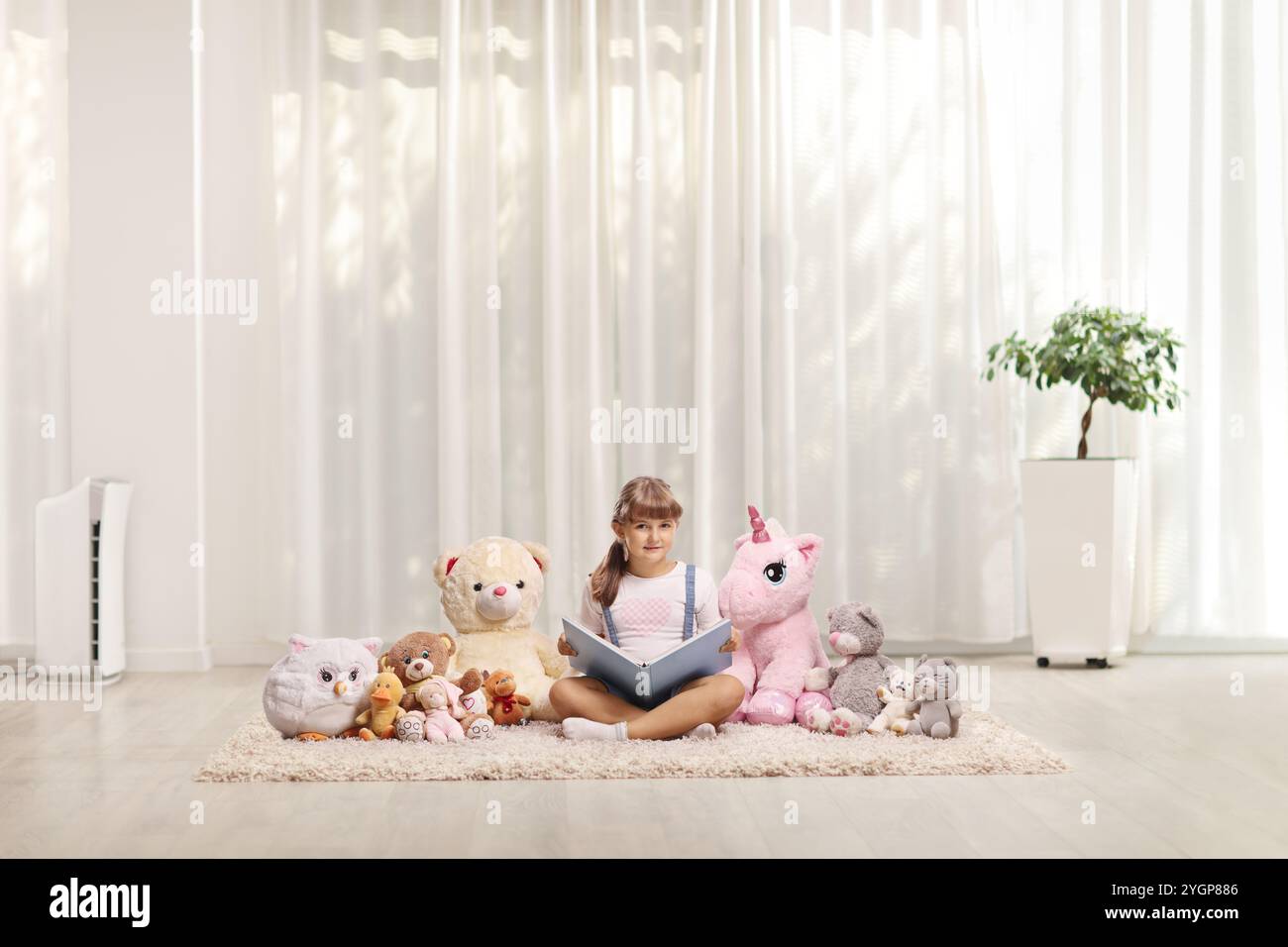 Little girl sitting on a carpet and reading a book to her plush animal ...