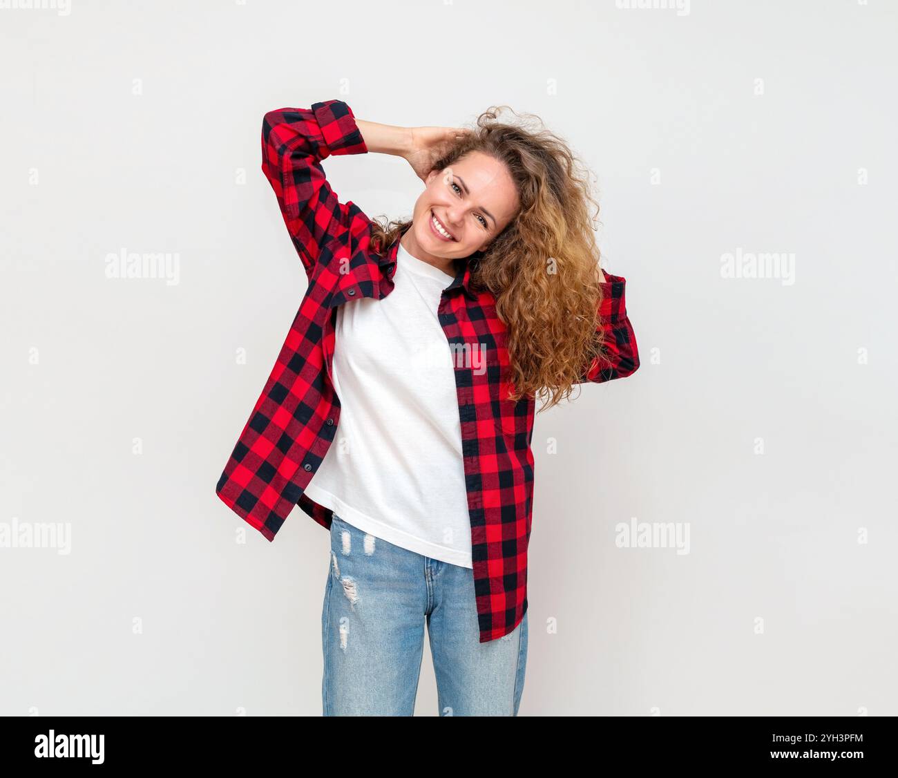 Cheerful curly woman in casual attire playfully poses for the camera in studio, holding hands behind head with positive emotions, carefree. Stock Photo