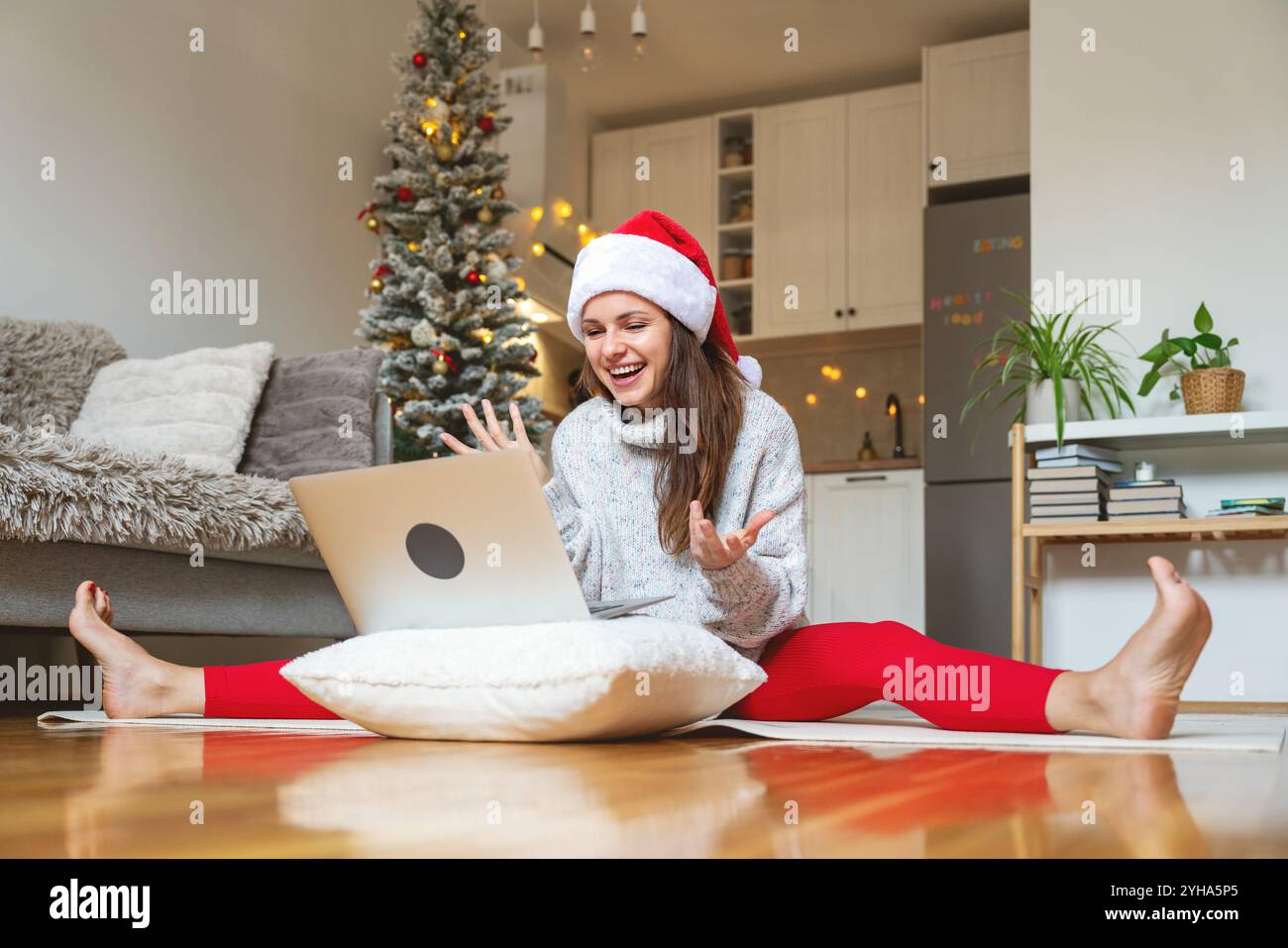 Woman in Santa hat and red pants sits on the floor in a split pose at home, using laptop for video call with friends or family during Christmas holida Stock Photo