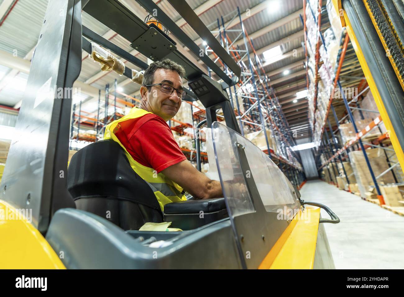 Happy manual worker turning to smile while driving forklift in distribution warehouse Stock Photo