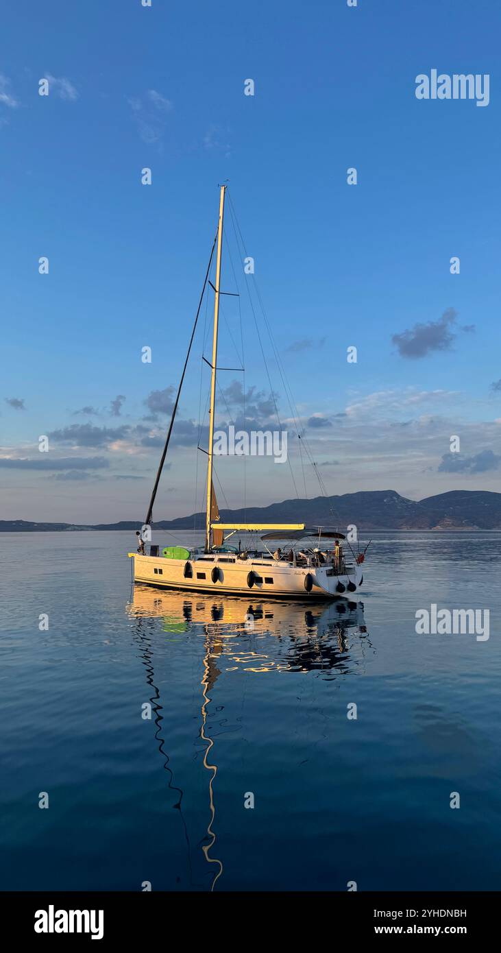 Modern sailing yacht anchored in calm Mediterranean waters at dusk reflects perfectly in mirror-like sea, with coastal mountains as backdrop Stock Photo