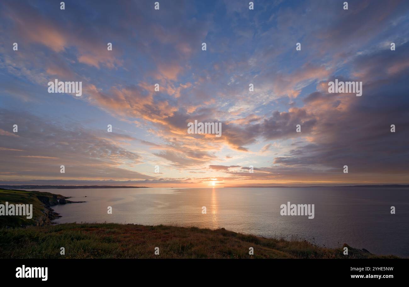 A beautiful sunset casts a warm glow over the tranquil ocean, with dramatic clouds and scenic cliffs in the foreground Stock Photo