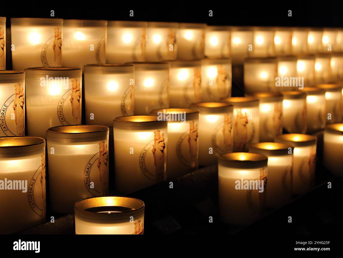 A row of lit candles in glass holders, casting a warm glow in a dimly lit space, evoking a peaceful, spiritual ambiance. Stock Photo