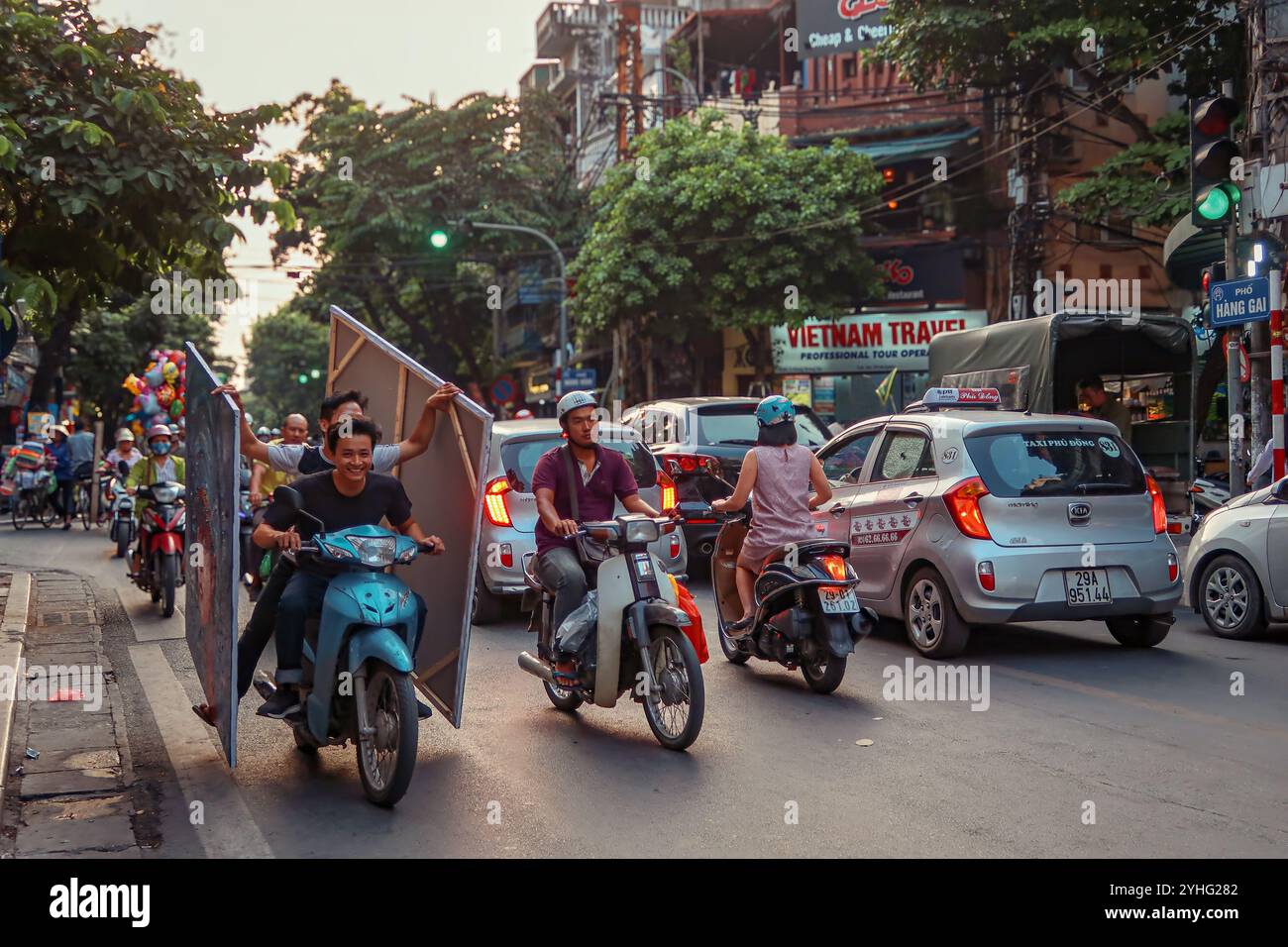 Motorcyclists carrying large, unwieldy items navigate through the busy streets of Hanoi, Vietnam, highlighting the daily hustle in the city's bustling Stock Photo