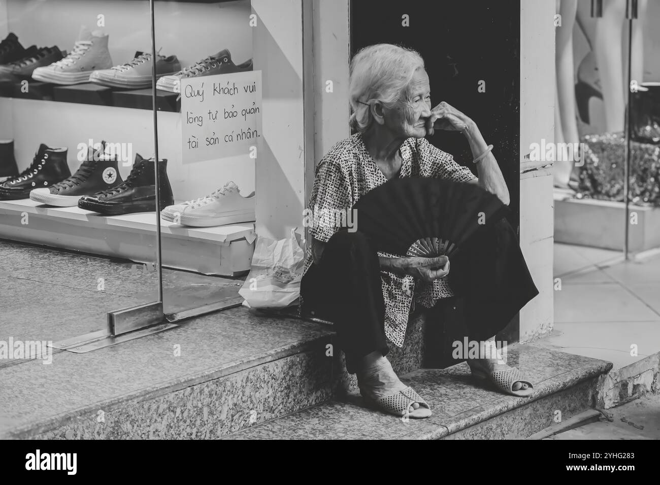 An elderly woman, seated on the steps of a store with a fan in hand, takes a moment of rest while the hustle of the street continues around her. Stock Photo