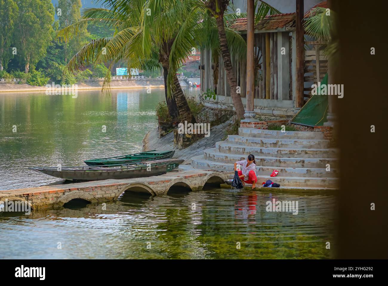 A serene riverside scene with a woman washing clothes, sitting by the steps near moored boats and tropical palms. Peaceful, rural life in Vietnam. Stock Photo