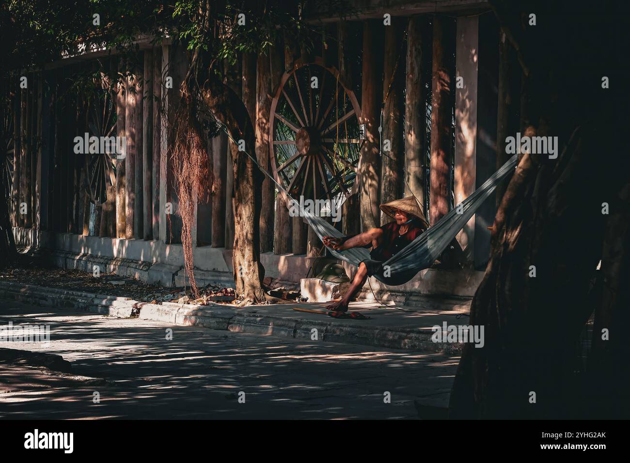A local man relaxes in a hammock, shaded under trees and wearing a traditional Vietnamese conical hat. The scene captures a quiet moment of daily life Stock Photo