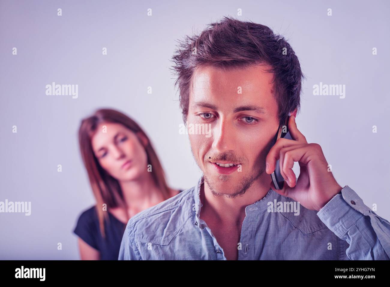 The man’s content expression while on a call contrasts with the woman’s blurred, watchful gaze. This symbolizes the complexities of trust and the subt Stock Photo