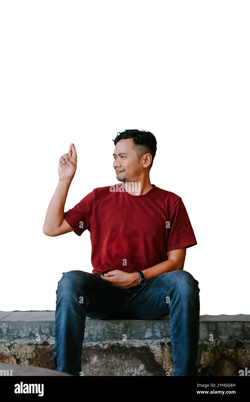 A man in a red T-shirt sits on a stone ledge, raising his hand with a serious expression against a plain white background, conveying confidence and fo Stock Photo