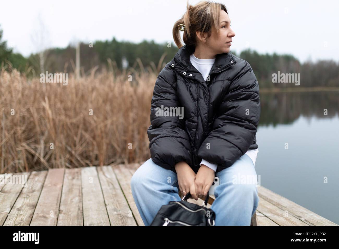 A woman sits on a wooden dock, looking out at a lake in the distance ...