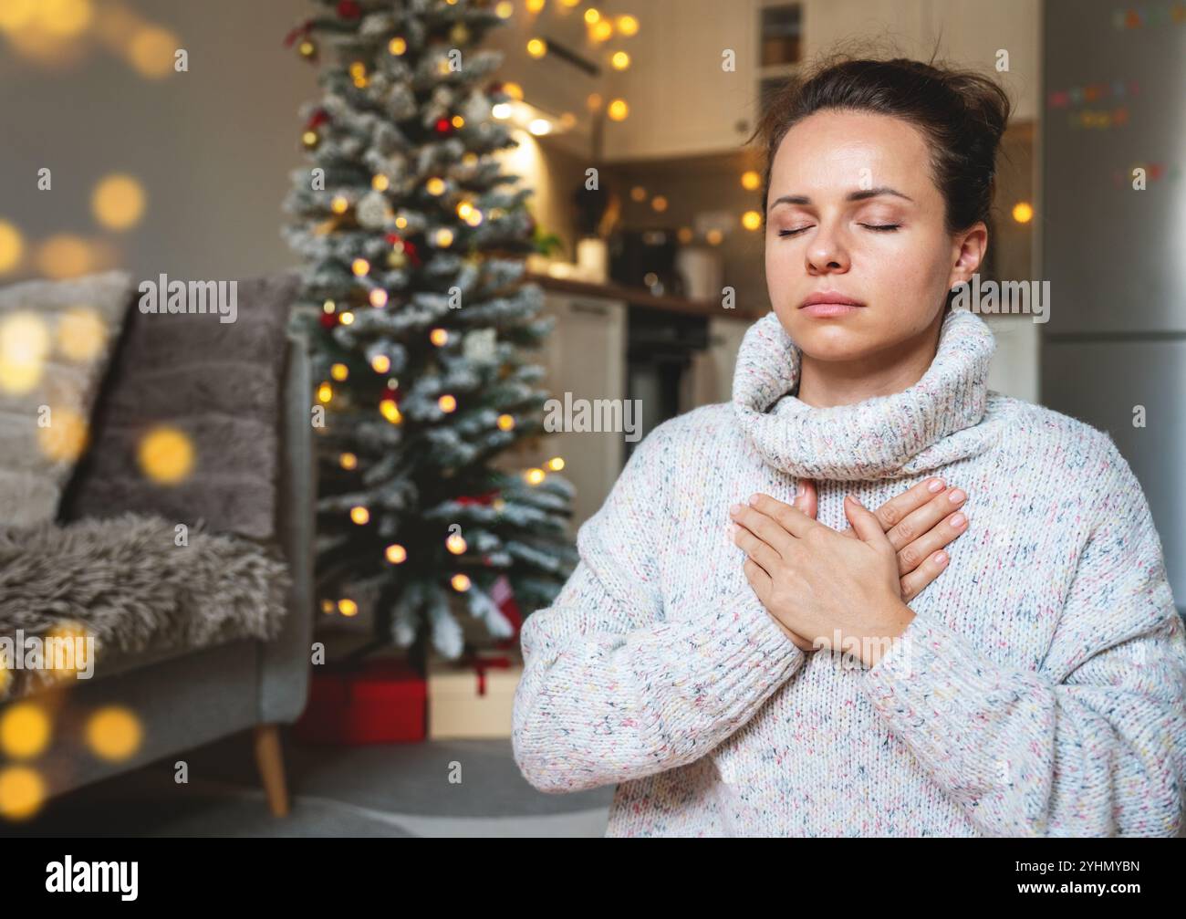 Woman in a warm knit sweater meditating and practicing breathing exercises at home during Christmas holidays, with a Christmas tree in the background. Stock Photo