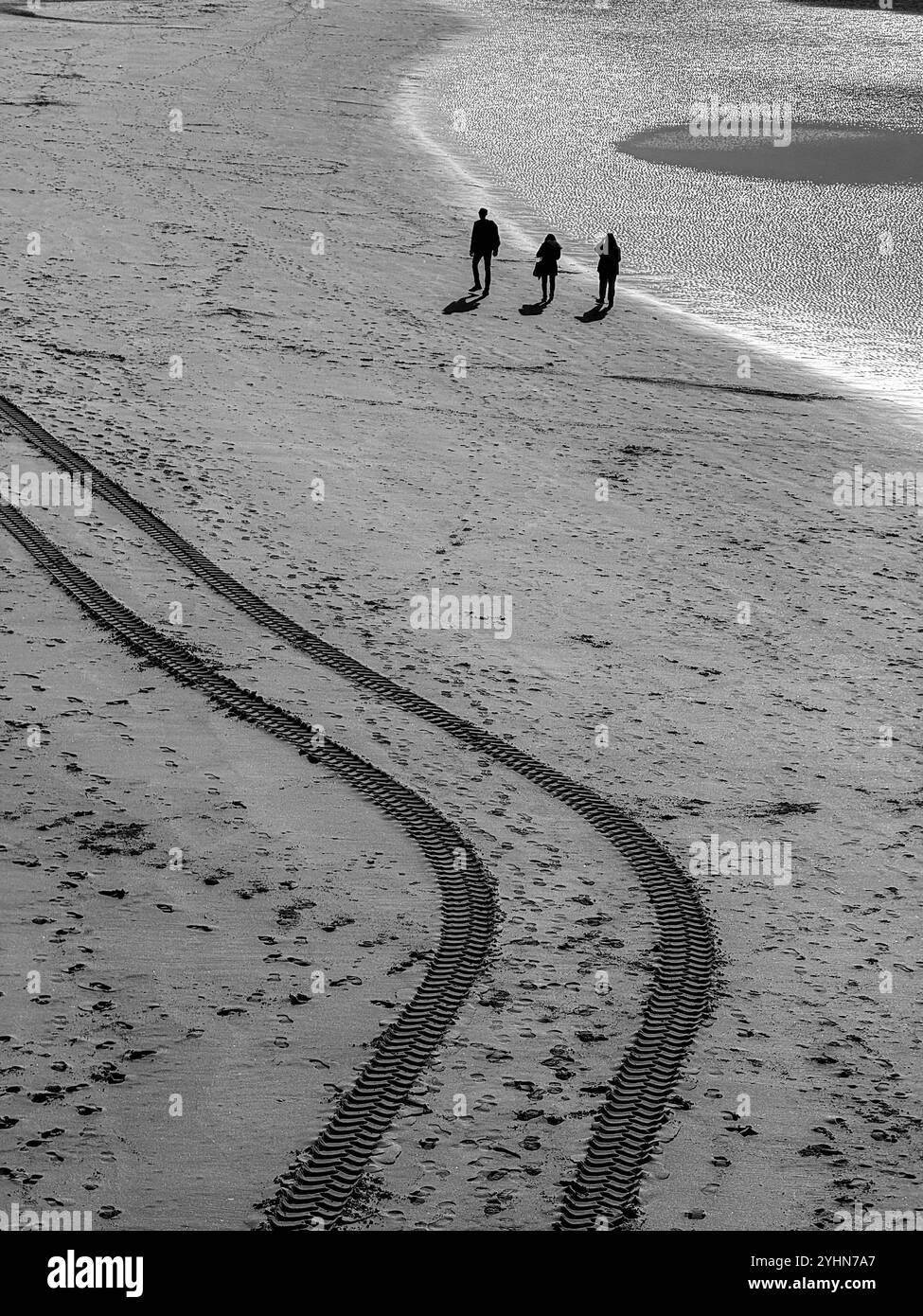 Three people on silhouette walking along beach at low tide besides fresh tyre tracks in the sand. Blackpool, Lancashire UK - Smartphone Captured Stock Image