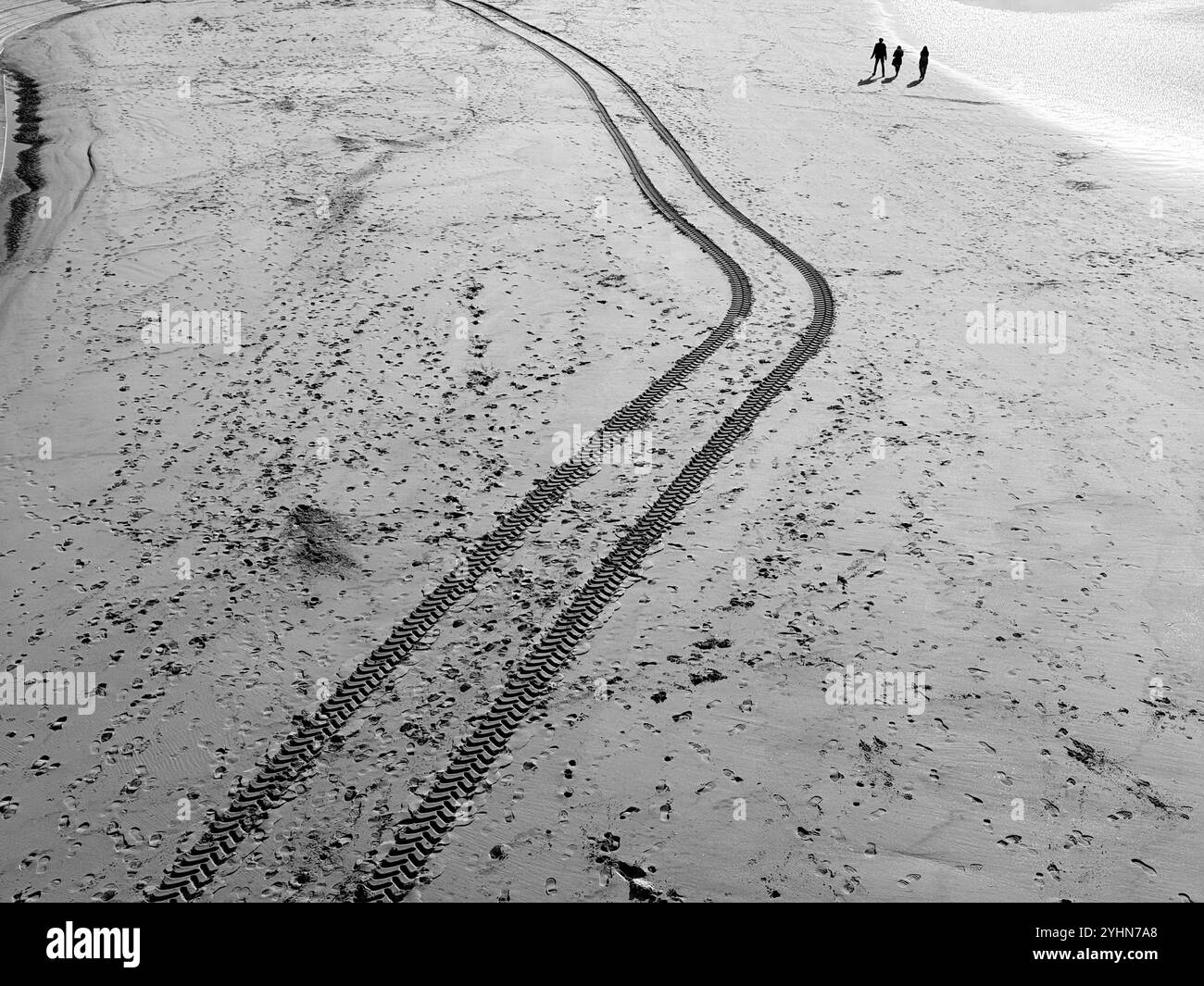 Three people on silhouette walking along beach at low tide besides fresh tyre tracks in the sand. Blackpool, Lancashire UK - Smartphone Captured Stock Image