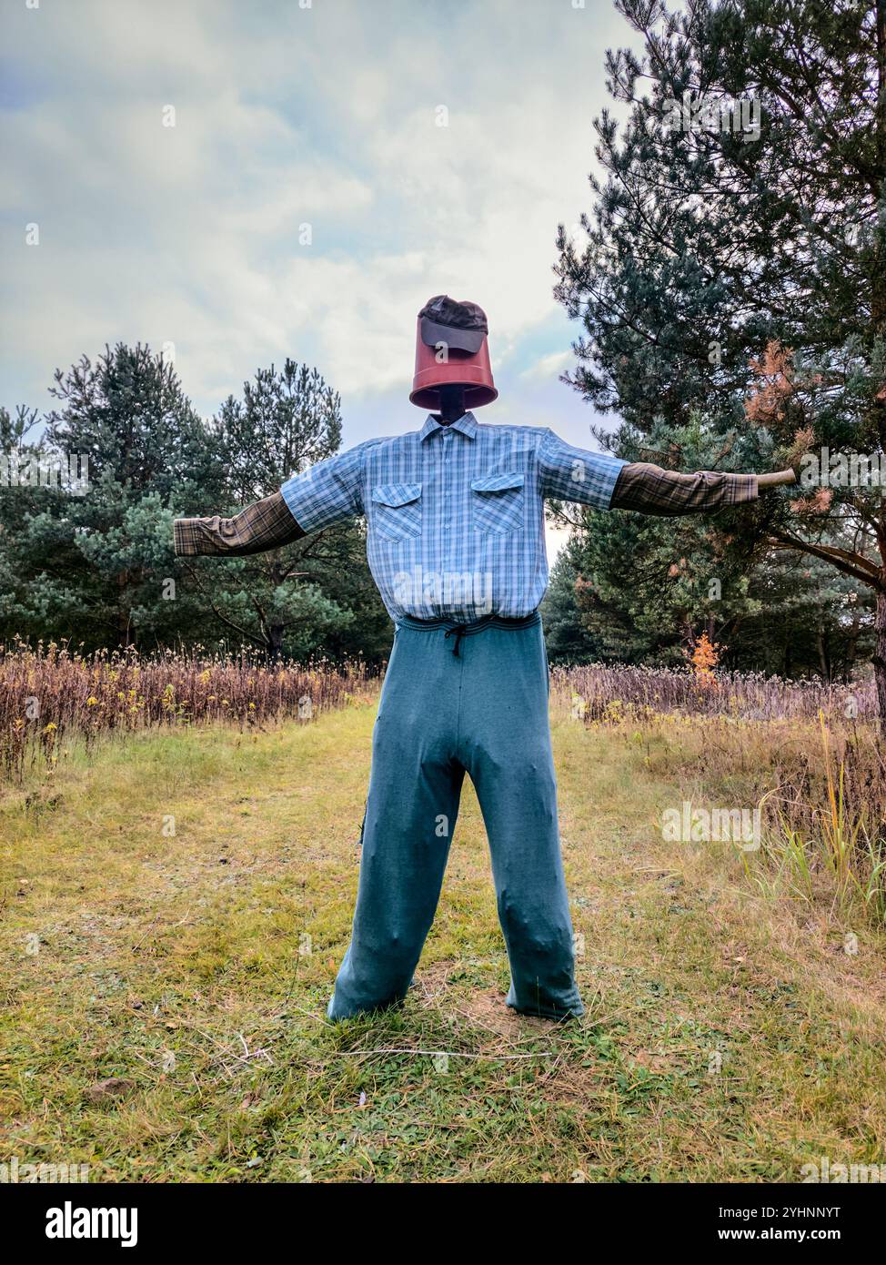Homemade scarecrow with a plastic pot for a head, wearing a plaid shirt, patched blue pants, and outstretched arms in a dirt road. - Smartphone Captured Stock Image