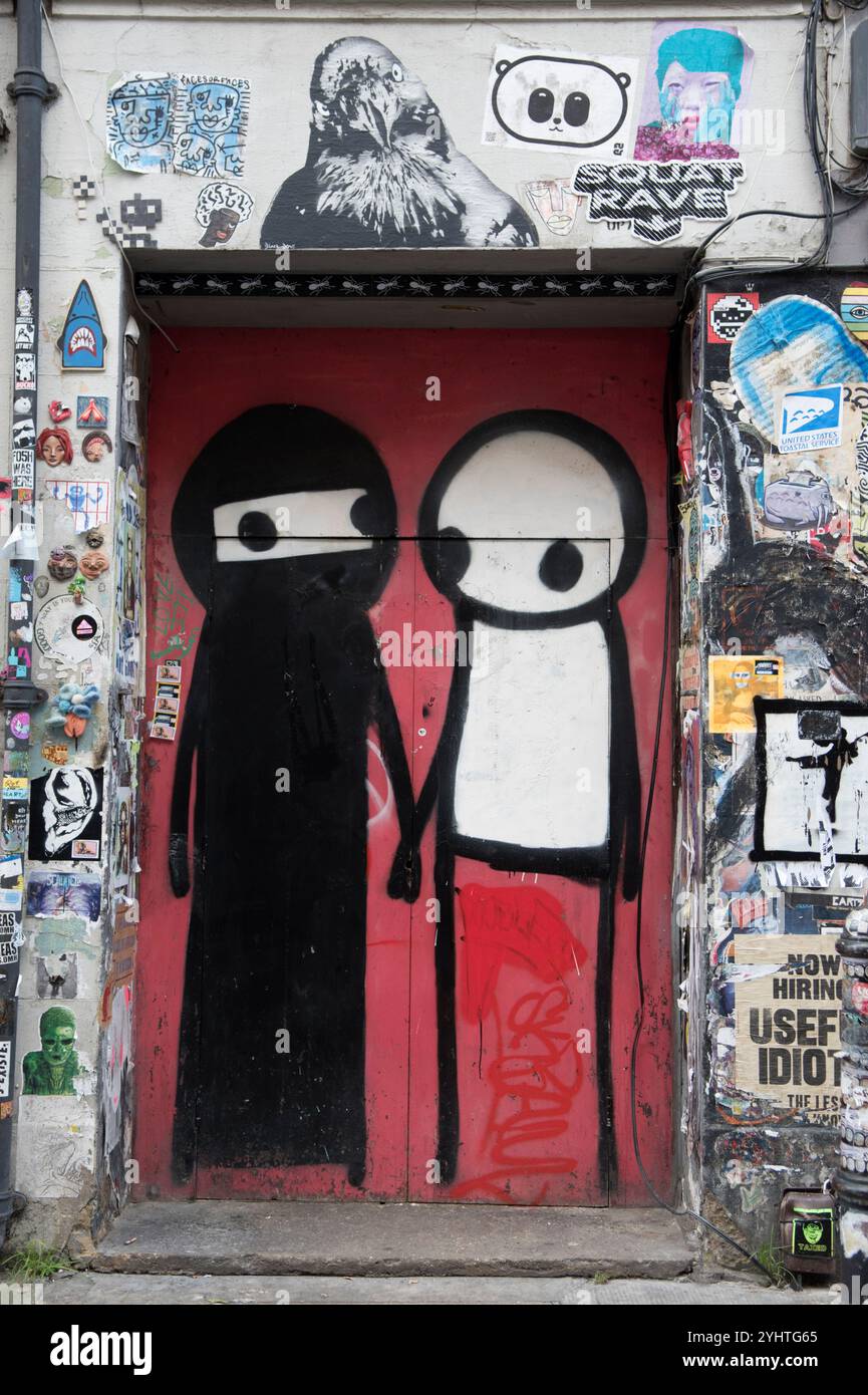 STIK graffiti street artist, wall art playfully depicting two Muslim women one wearing her traditional black Abaya and Niqab and the other with no face covering or a white face covering allowing just the eyes to see. Just off Brick Lane, a club doorway surrounded by graffiti.  Princelet Street,Tower Hamlets, Spitalfields, East End London England UK 2020s 2024 HOMER SYKES Stock Photo