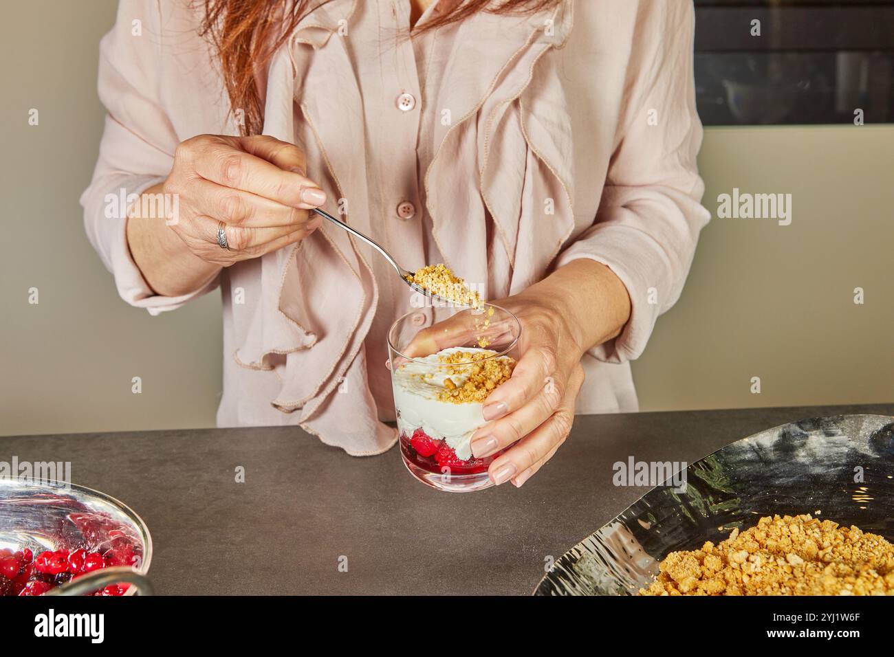 A person adds a crumb layer to a parfait in a glass, creating a crunchy texture to complement the creamy and fruity layers. Stock Photo