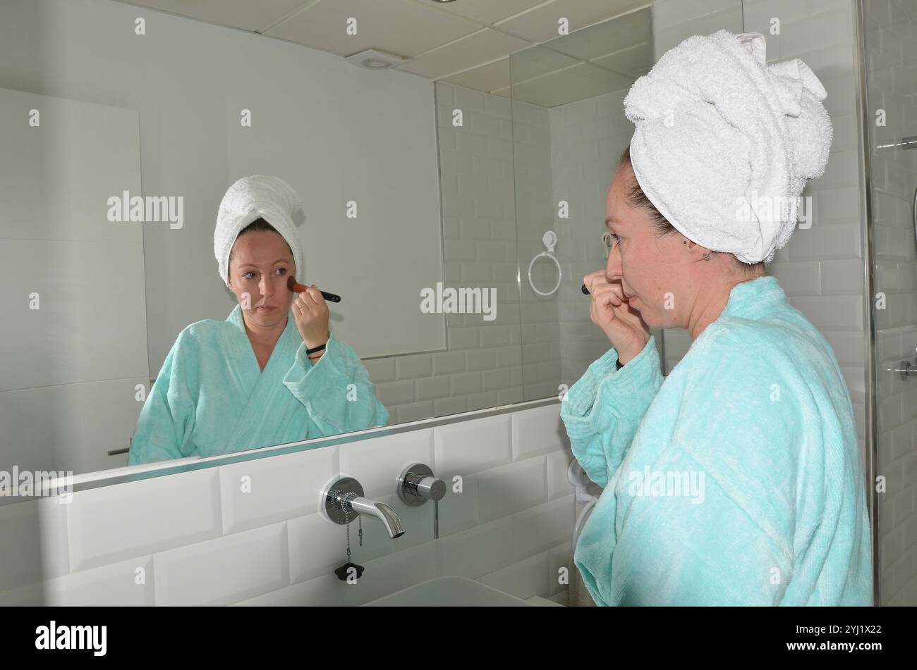 Woman applying makeup with brush in bathroom mirror wearing bathrobe and towel turban Stock Photo