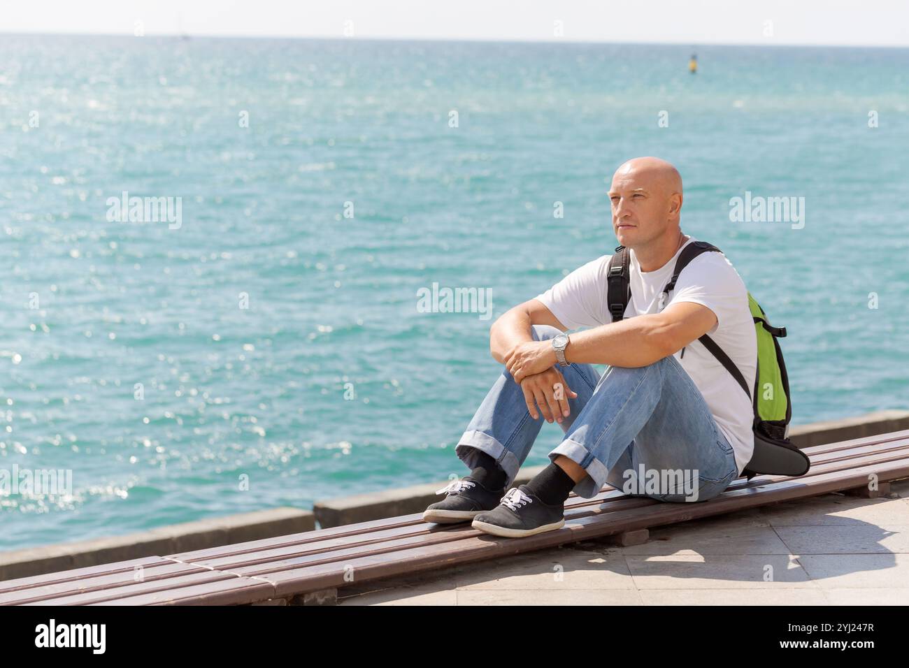 Man sits on a bench by the ocean, looking out at the water. He is wearing a backpack and a white shirt. The scene is peaceful and calming, with the so Stock Photo