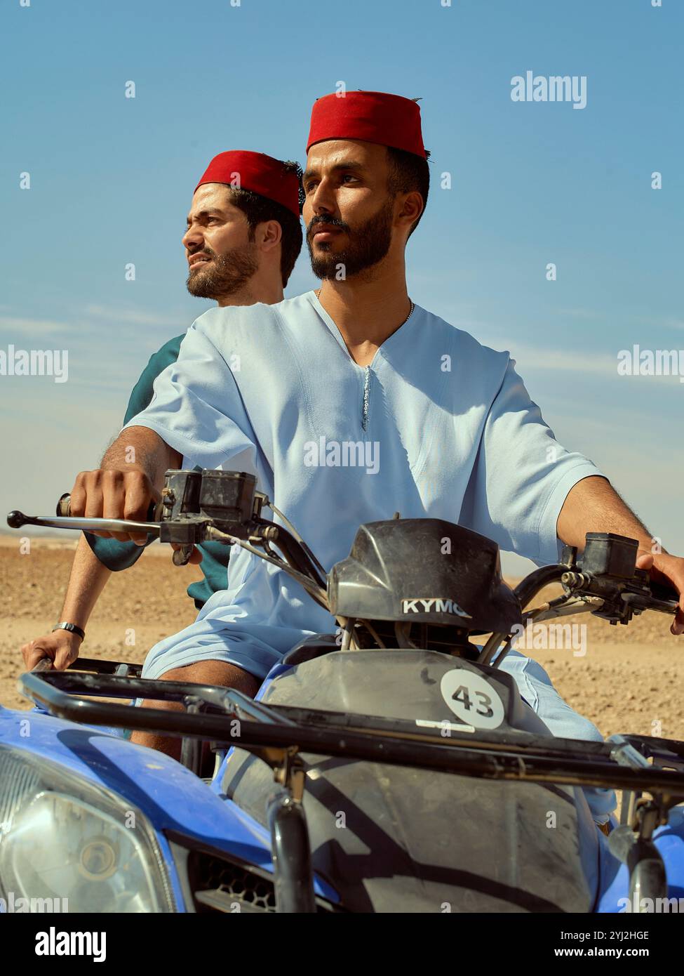 Two men in traditional attire riding a motorcycle through a desert landscape, Morocco Stock Photo