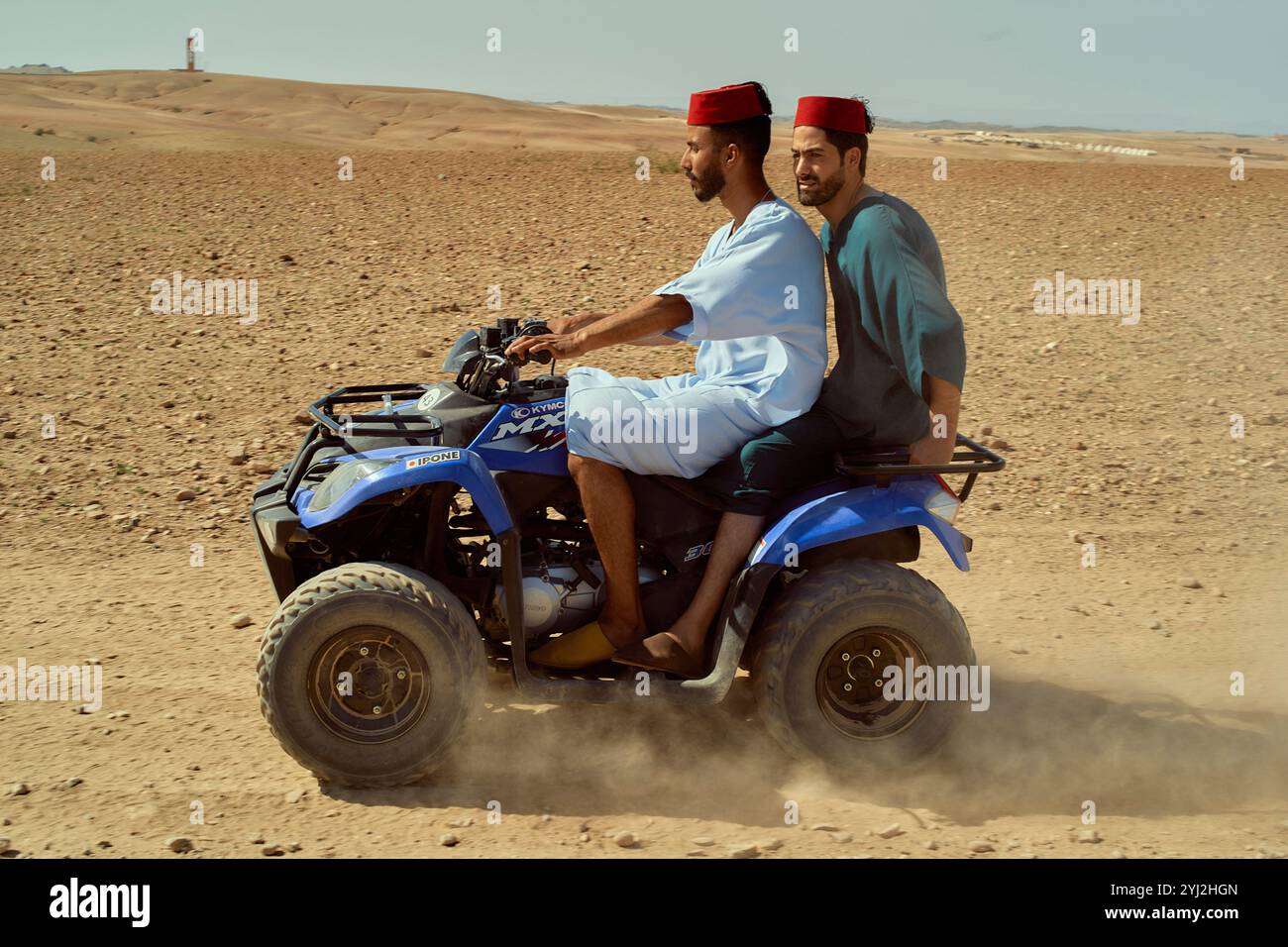 Two men in traditional attire riding a blue ATV in a desert landscape, Morocco Stock Photo