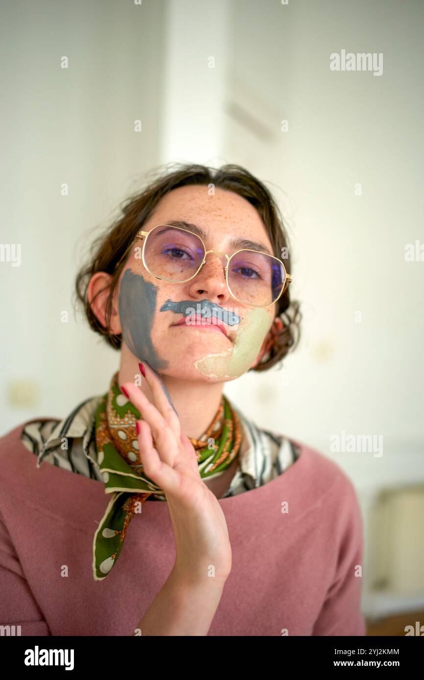 Young woman with a painted face and glasses posing thoughtfully in a bright room. Stock Photo