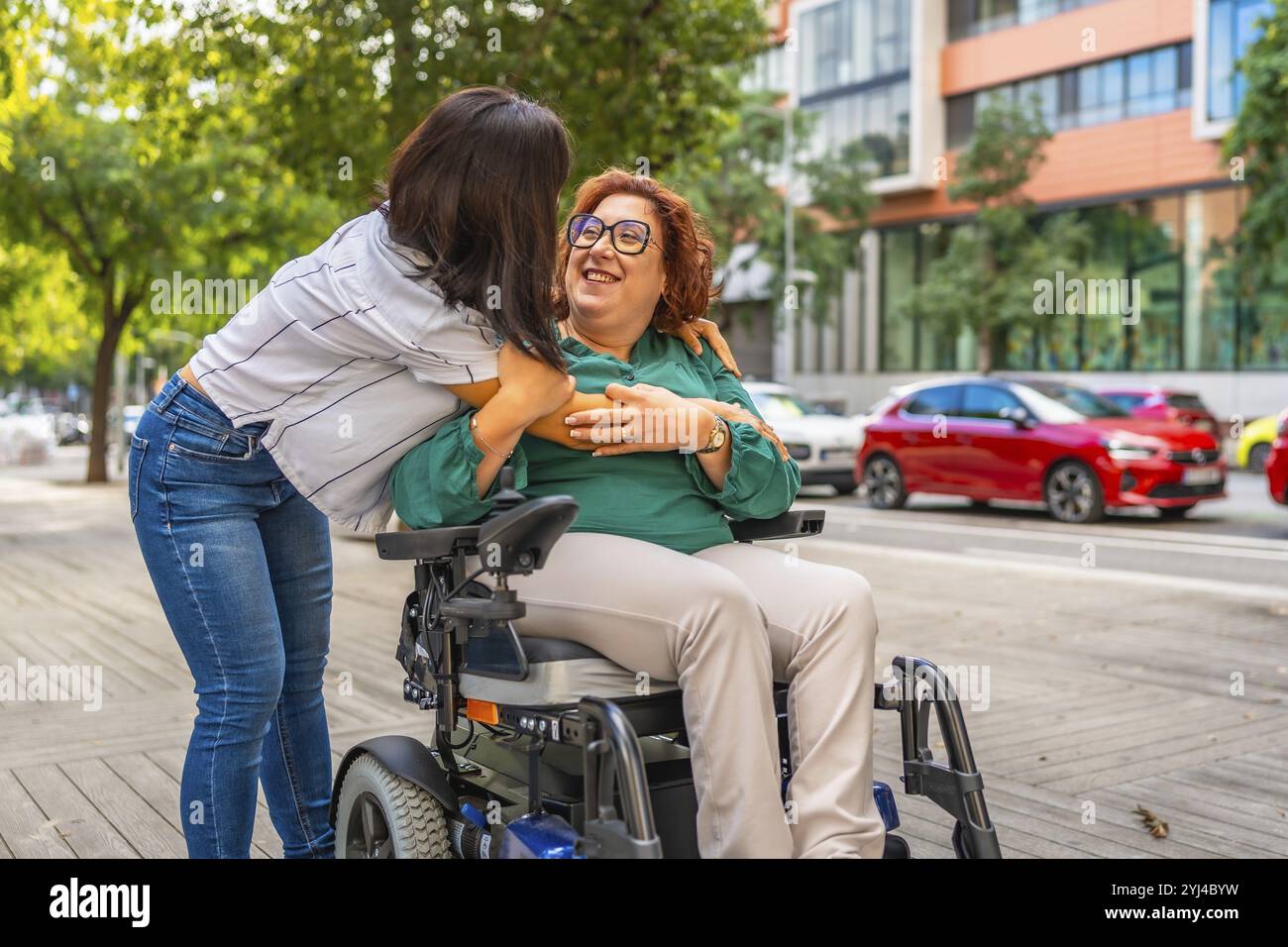 Happy adult caucasian disabled woman in electric wheelchair and asian female friend hugging in the street Stock Photo