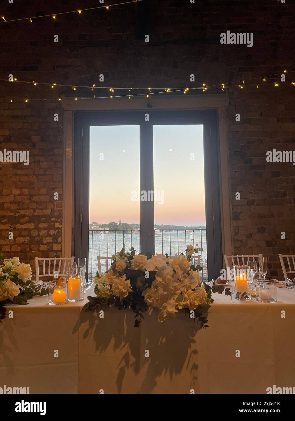 A romantic wedding table setting with flowers, candles, and fairy lights, framed by a sunset through the window. - Smartphone Captured Stock Image