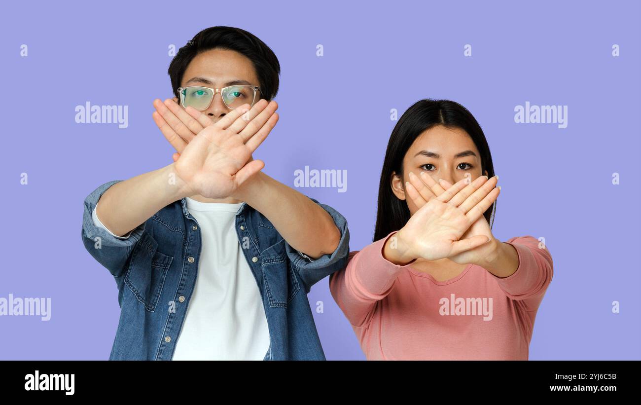 We don't talk. Young asian couple holding crossed palms against mouths, grey background Stock Photo