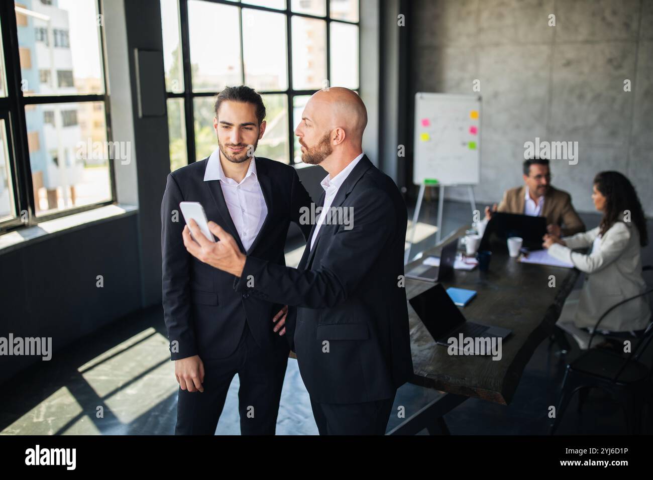 Two men in suits are engaged in a discussion while looking at a smartphone. In the background, colleagues are working at a large table in a stylish of Stock Photo