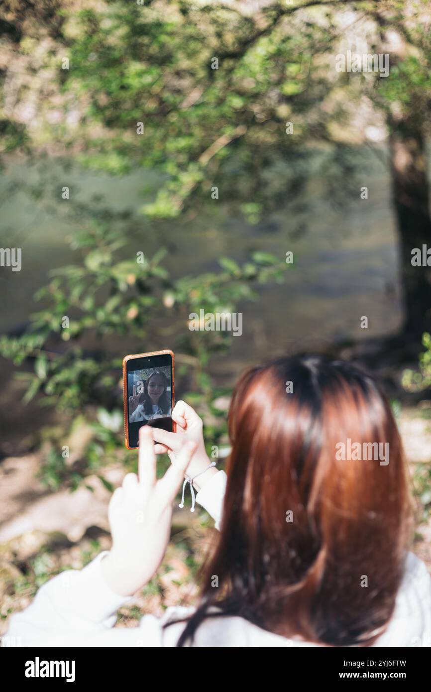 A woman with red hair is seen from behind, taking a selfie with her smartphone while standing near a river in a forested area. Stock Photo