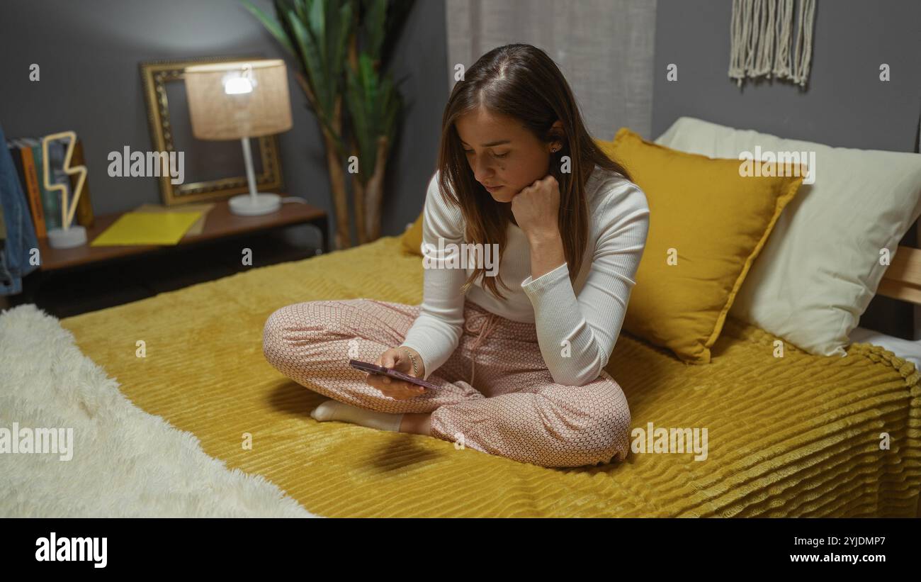 A young, attractive, blonde, caucasian woman sits thoughtfully on a bed in a cozy, indoor bedroom at home, using her phone. Stock Photo