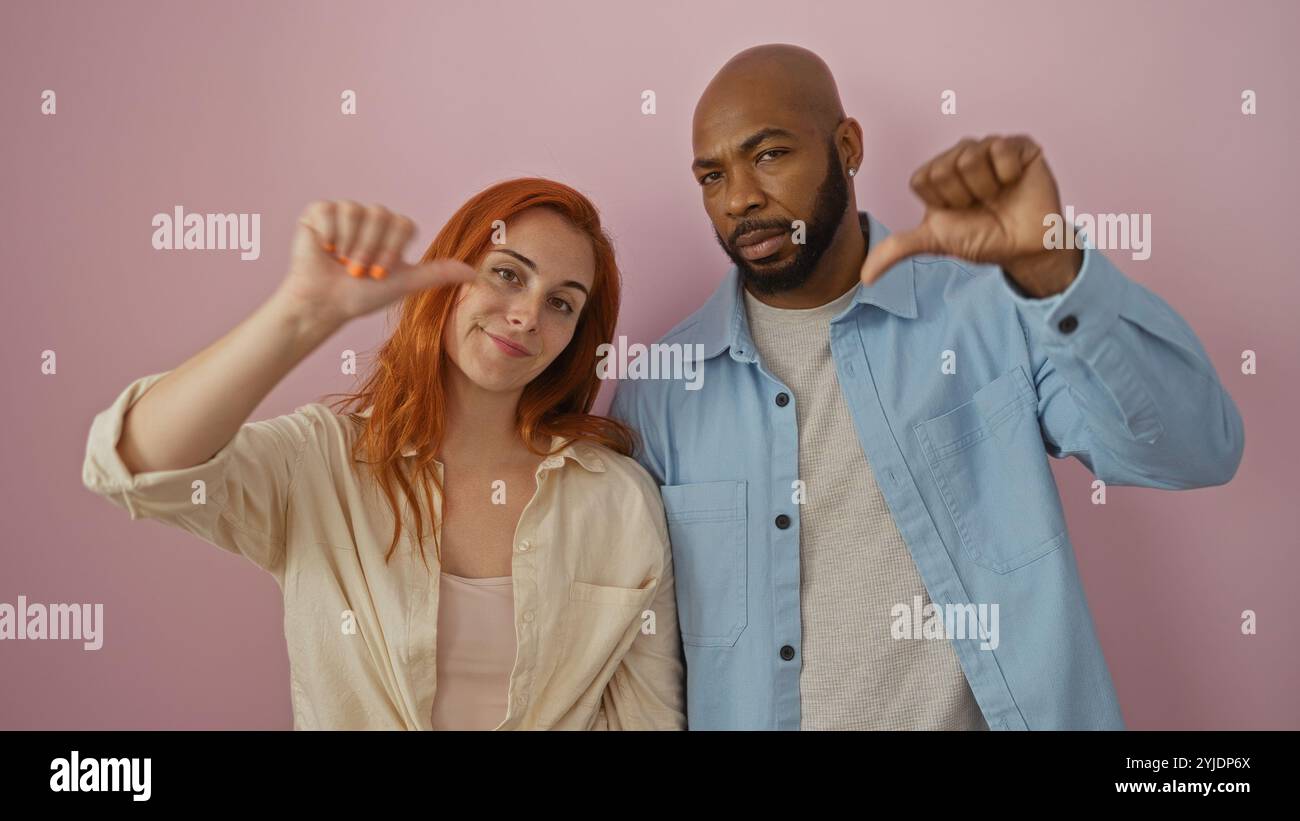Interracial couple, man and woman, standing together against a pink background, showing thumbs down, expressing disapproval or dissatisfaction, with b Stock Photo