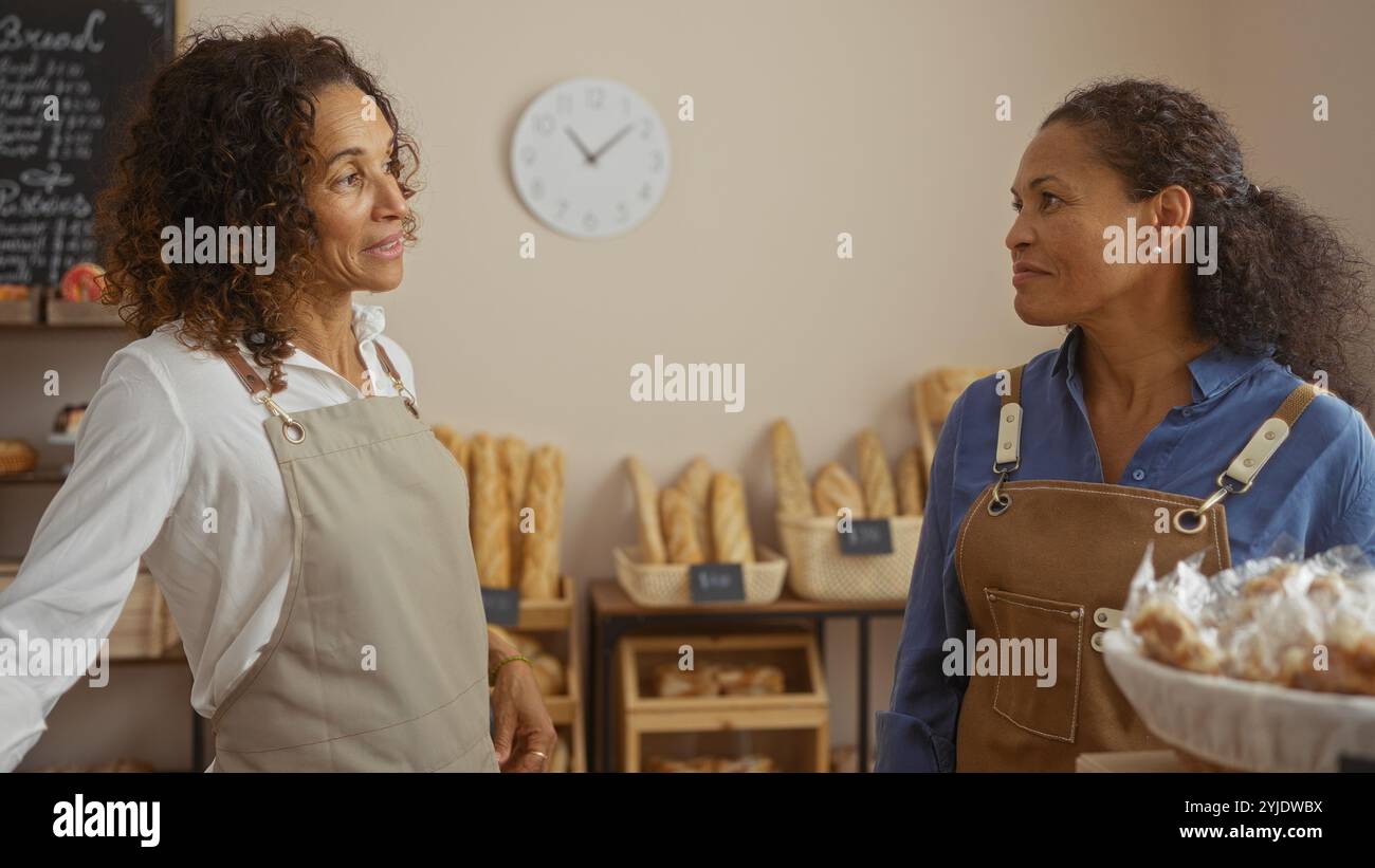 Women bakers discussing work in a cozy bakery shop with shelves of fresh bread in the background and a clock on the wall Stock Photo