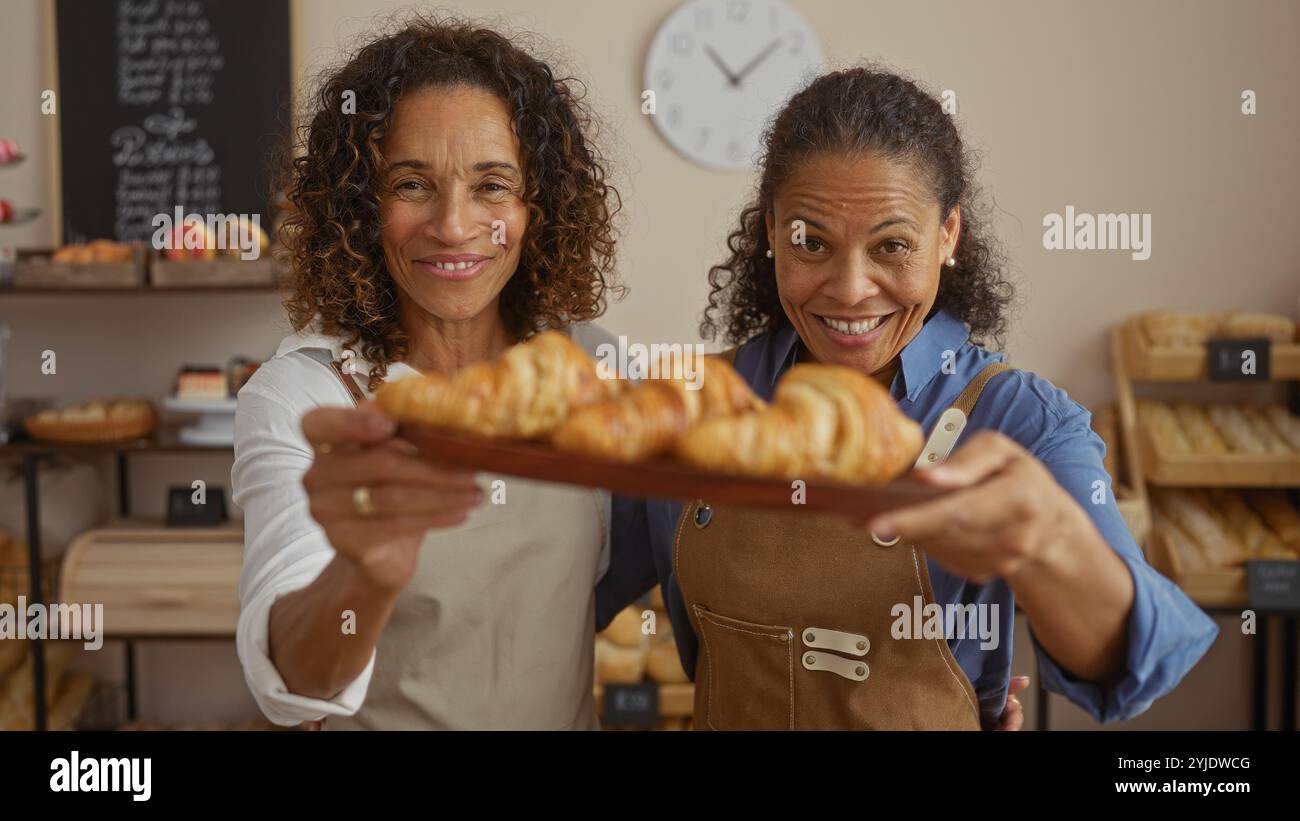 Women bakers smiling in bakery holding tray of freshly baked croissants in cheerful, inviting shop interior featuring bread shelves and wall clock in Stock Photo