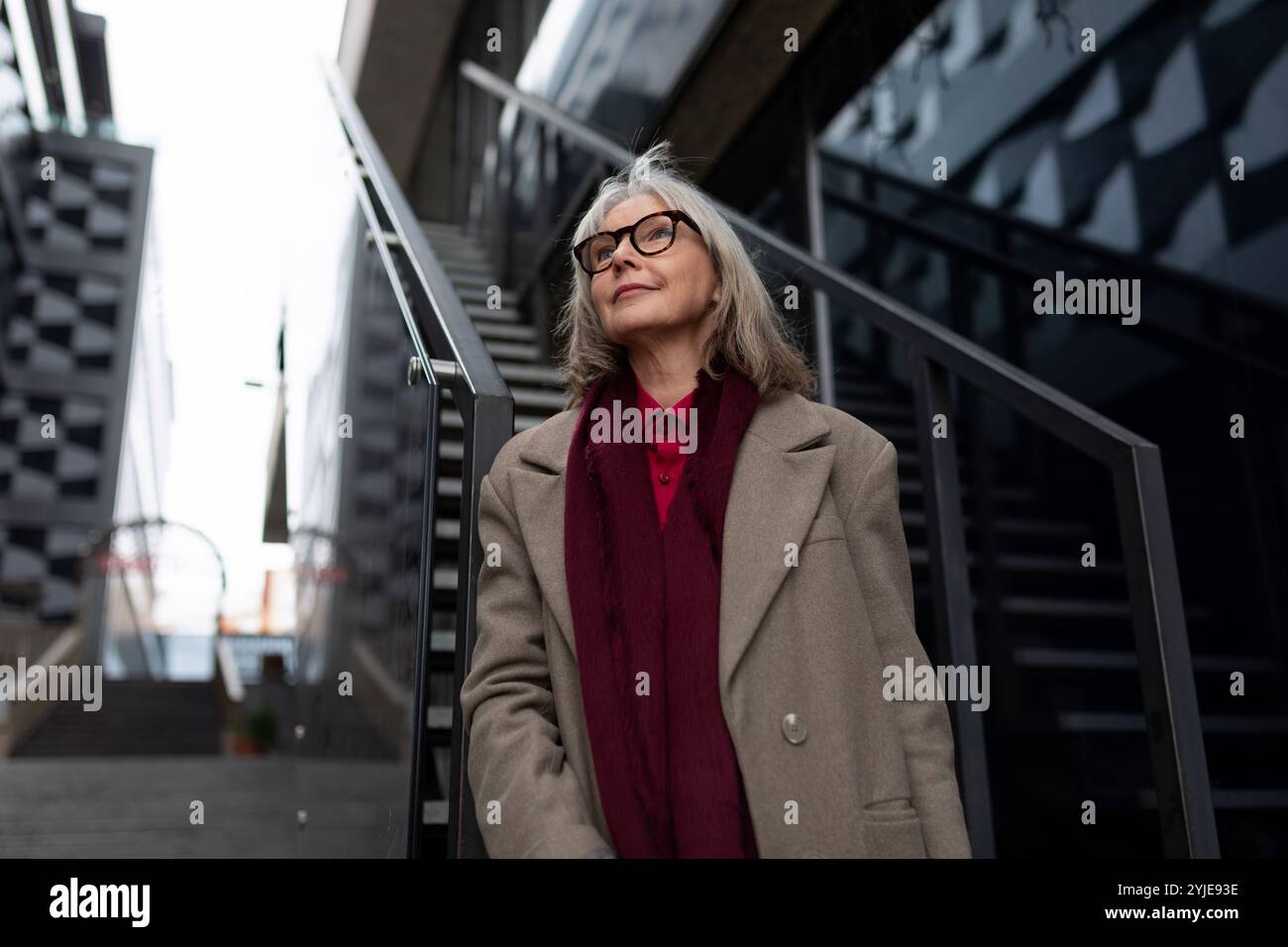 An older woman wearing glasses and a coat stands on steps between ...