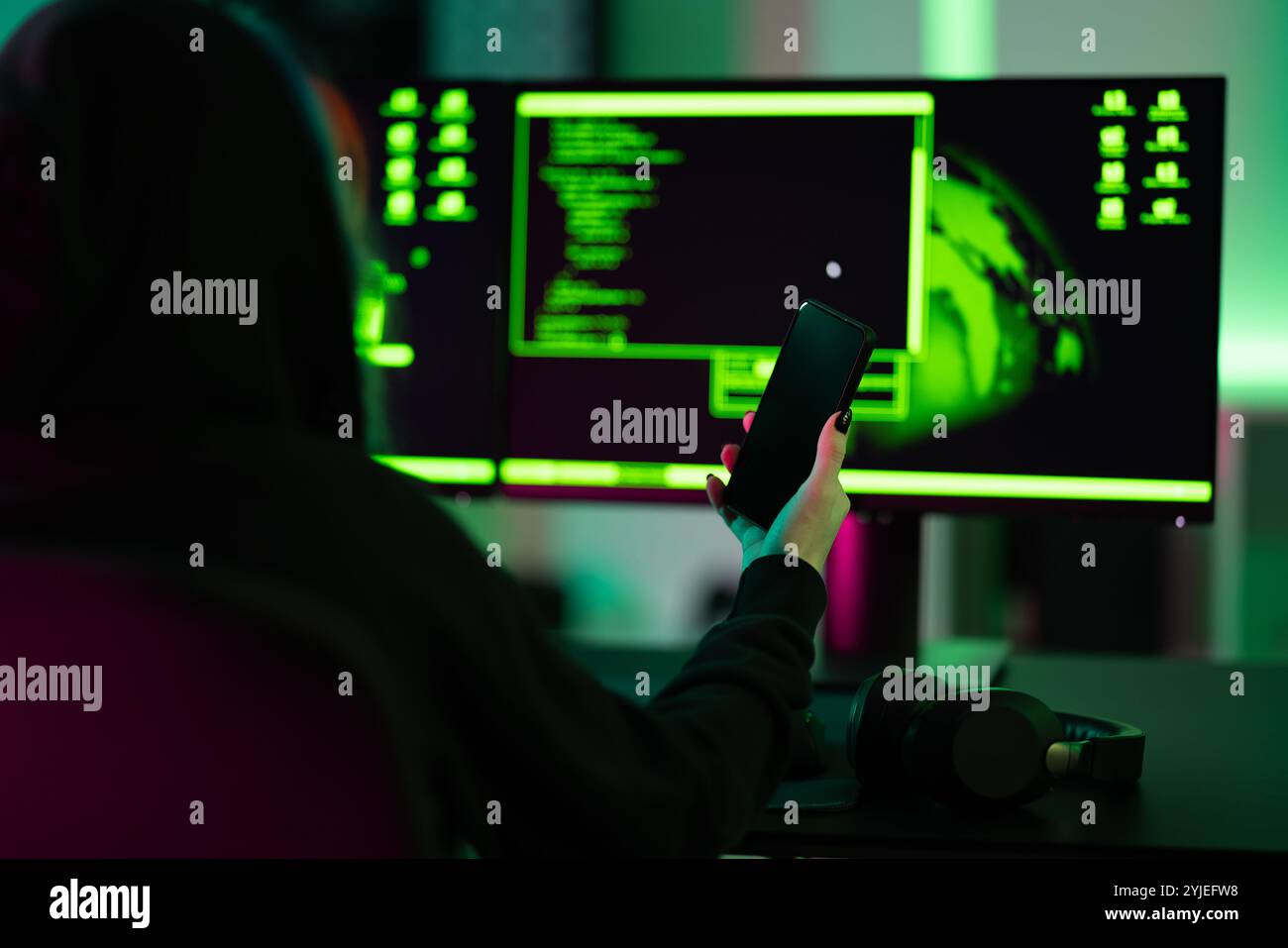 Hacker holding smartphone in front of dual screens with coding interface Stock Photo