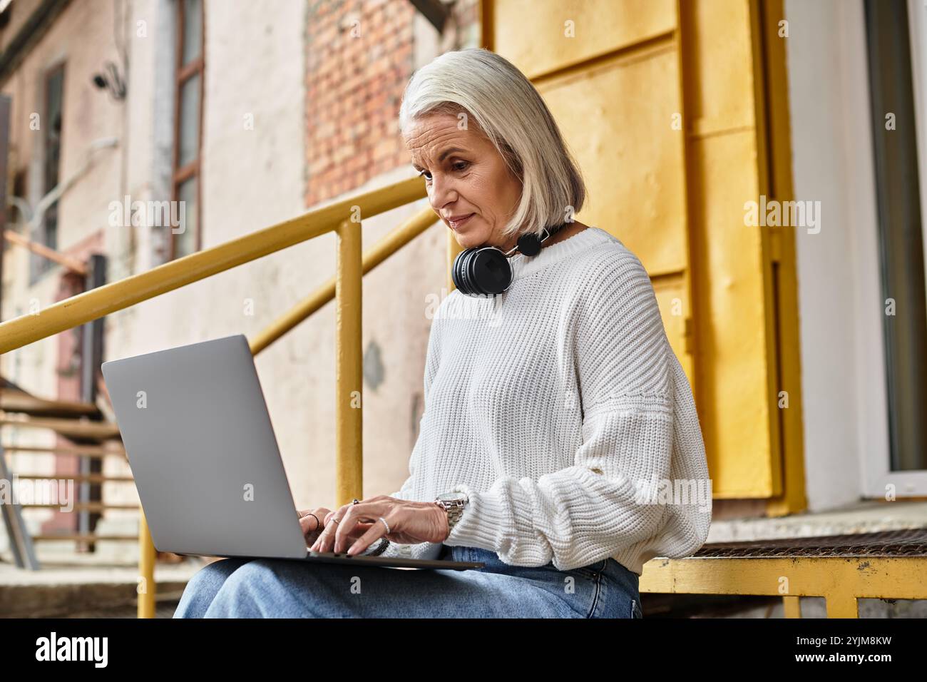 A mature woman with gray hair sits on steps, focused on her laptop and ...